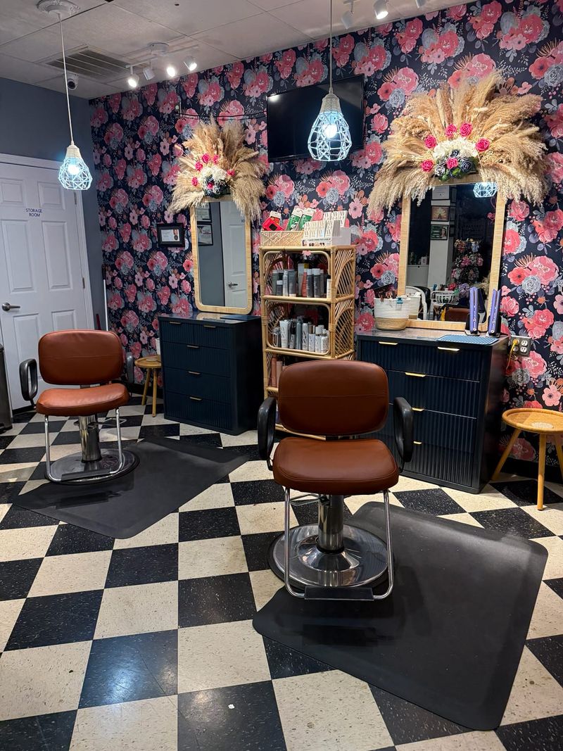 Two styling chairs on a black and white checkerboard floor, facing mirrors set into dark floral wallpaper, with pampas grass above them.